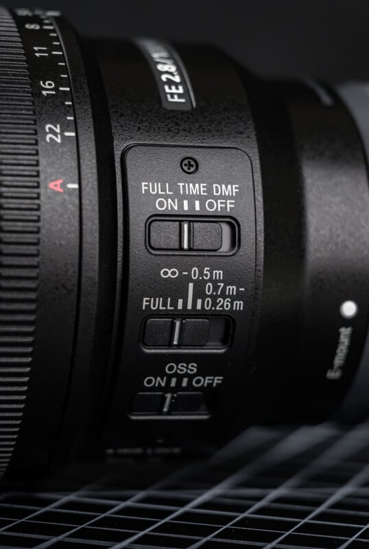 Close-up of a camera lens showing switches for full-time DMF, focusing distance (infinity to 0.26m), and OSS (optical steady shot) with grid lines beneath the lens.