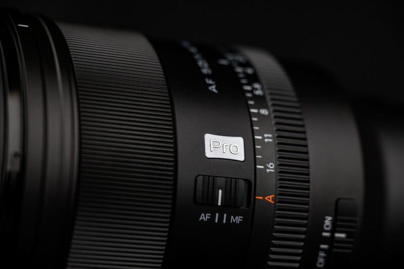 Close-up of a professional camera lens showing a "Pro" label, focus mode switch with AF and MF options, and an aperture ring with numbers and an "A" marking. The lens appears black with textured and smooth surfaces.