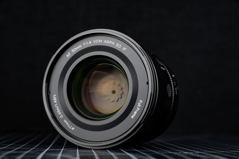 Close-up of a camera lens with "AF 50mm 1:1.4 VCM ASPH ED IF" labeled on the front, placed on a grid-patterned surface against a dark, blurred background.