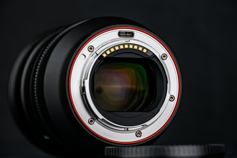 Close-up of a camera lens mount with gold electronic contacts, a red ring around the edge, and reflections visible on the lens glass against a dark background.