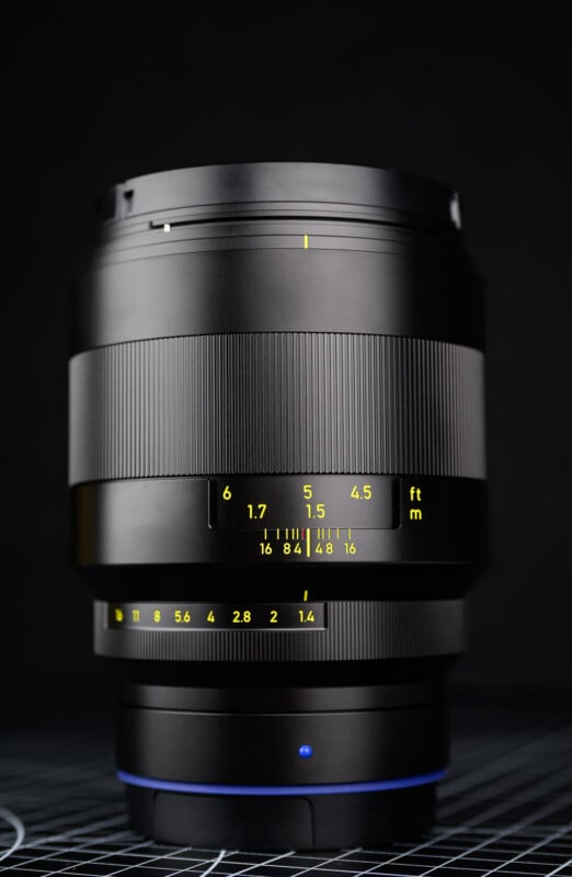 A close-up photo of a black camera lens with yellow distance and aperture markings, set against a dark background. The lens stands upright on a grid-patterned surface.