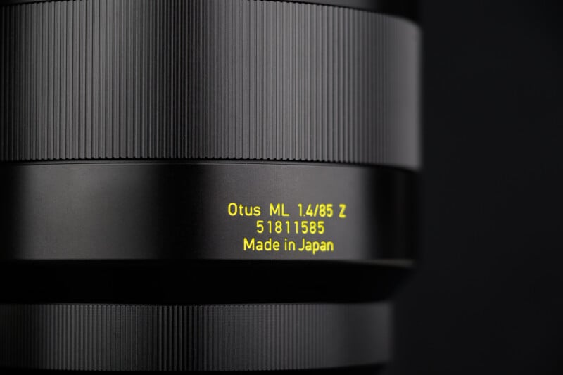 Close-up of a camera lens with the text " Otus ML 1.4/85 Z 51811585 Made in Japan" printed in yellow on the black lens barrel against a dark background.