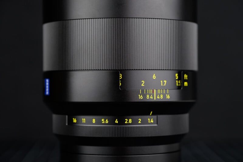 Close-up of a black camera lens showing detailed focus and aperture markings in yellow, with sharp grooves on the focus ring and a blurred dark background.