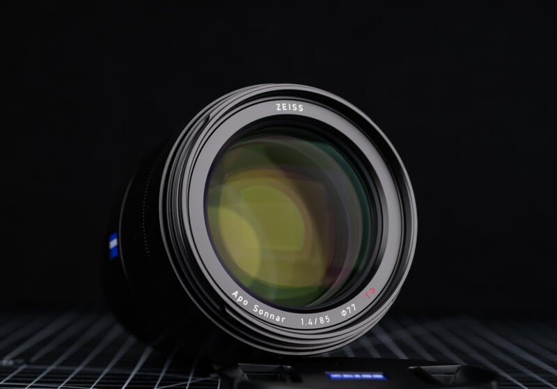 A close-up of a Zeiss camera lens, Apo Sonnar 1.4/85, placed on a patterned surface with a lens cap nearby, set against a dark, blurred background.