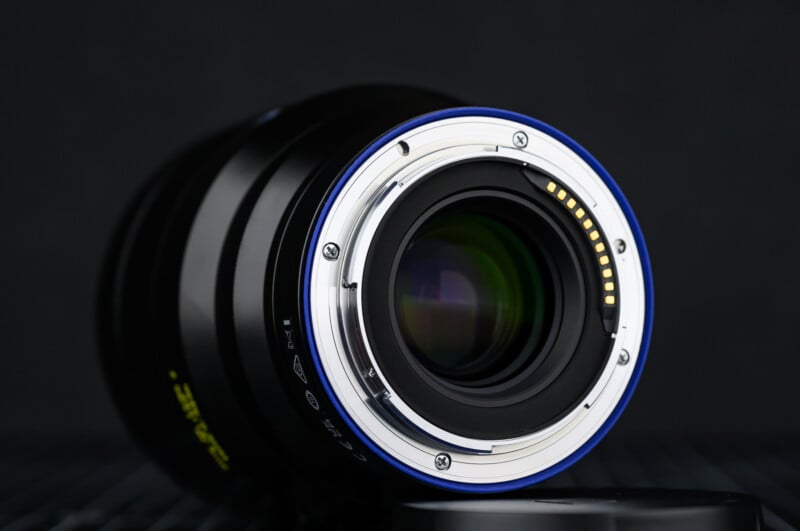 Close-up of the rear side of a camera lens showing the metal mount, electronic contacts, and glass elements, set against a dark, blurred background.