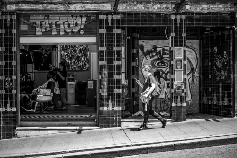 A person with a backpack walks past a tattoo shop on a sloped street; a person sits inside the shop while another works, and graffiti covers the door and window. The scene is in black and white.