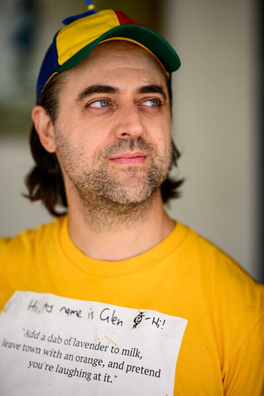 A man with long brown hair and light stubble wears a colorful cap and a yellow shirt with a handwritten name tag that says "Hi, my name is Glen" and a quote about lavender, milk, an orange, and laughter.