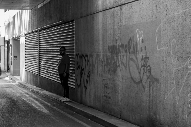 A man stands alone on a narrow sidewalk next to a graffiti-covered wall under an overpass in black and white; light filters in from the left, casting shadows along the alley.