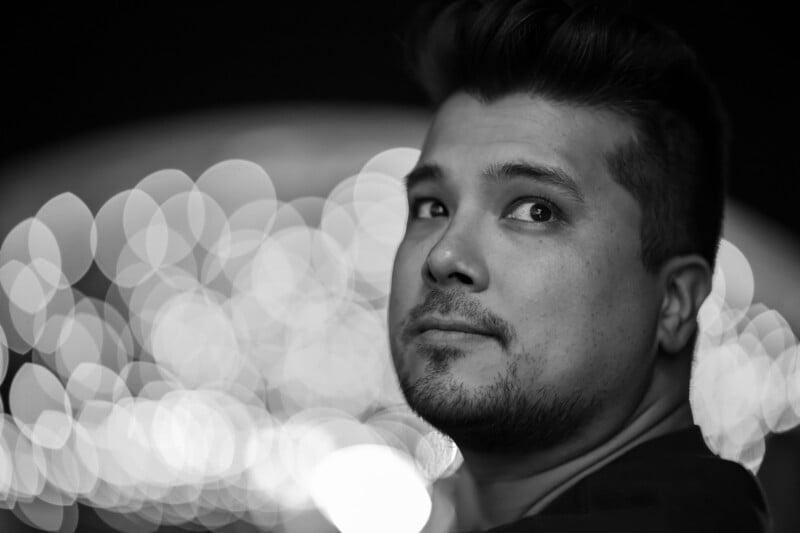 A man with short hair and slight facial hair looks over his shoulder at the camera. The background is blurred with large, circular bokeh lights, creating an artistic and dramatic effect. The photo is in black and white.