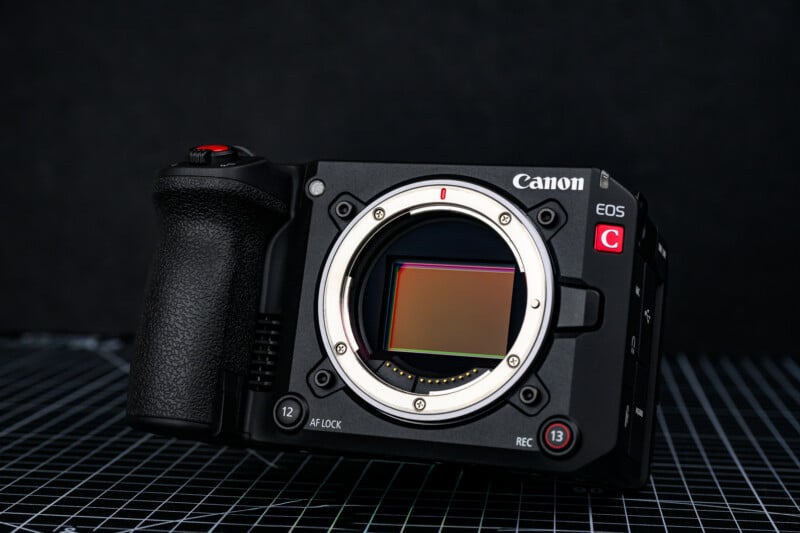 A Canon EOS cinema camera body is shown without a lens, revealing the sensor. The camera is black, positioned at an angle, and set against a dark, grid-patterned background.