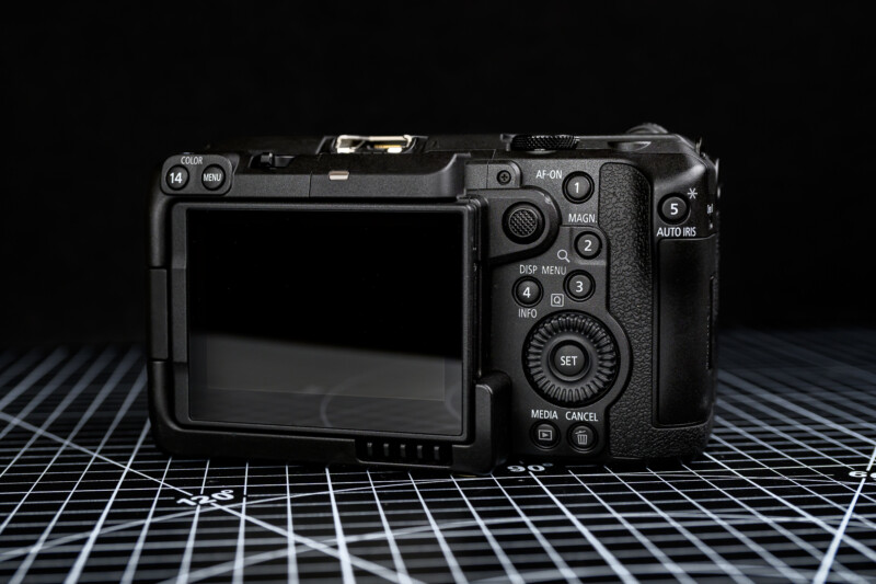 A digital camera shown from the back, displaying its screen and various buttons. The camera is placed on a black grid-patterned surface, with a dark background behind it.