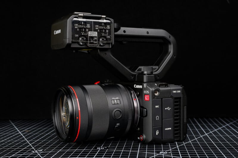 A Canon EOS cinema camera with a large lens, top handle, and audio interface is positioned on a black grid-patterned surface against a black background.
