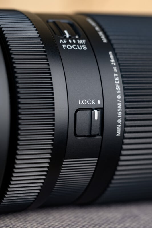 Close-up of a camera lens showing the AF/MF focus switch and a lock slider, with visible textured grip rings and detailed labeling against a soft, neutral background.