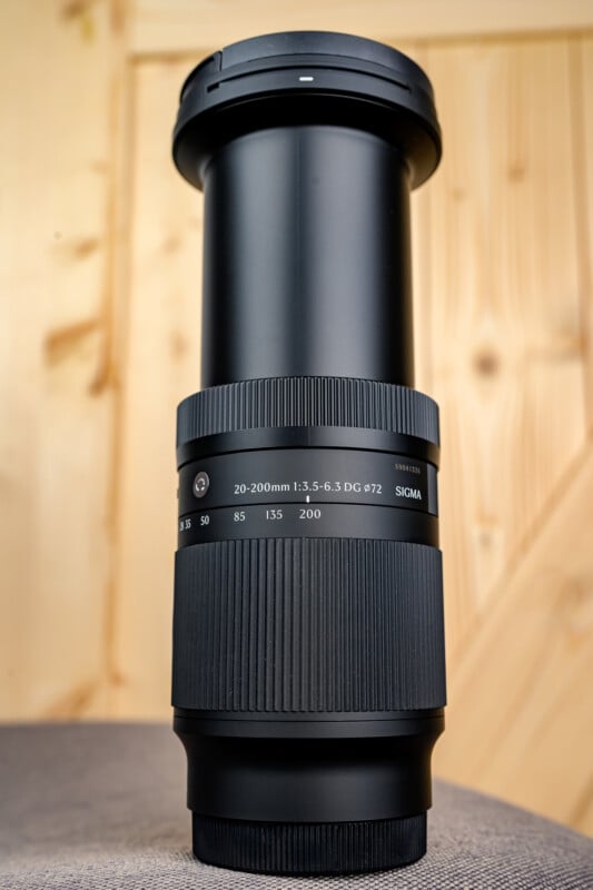 A Sigma 20-200mm camera zoom lens is standing upright on a gray surface with a wooden background. The lens is extended, showing its full length and textured grip.