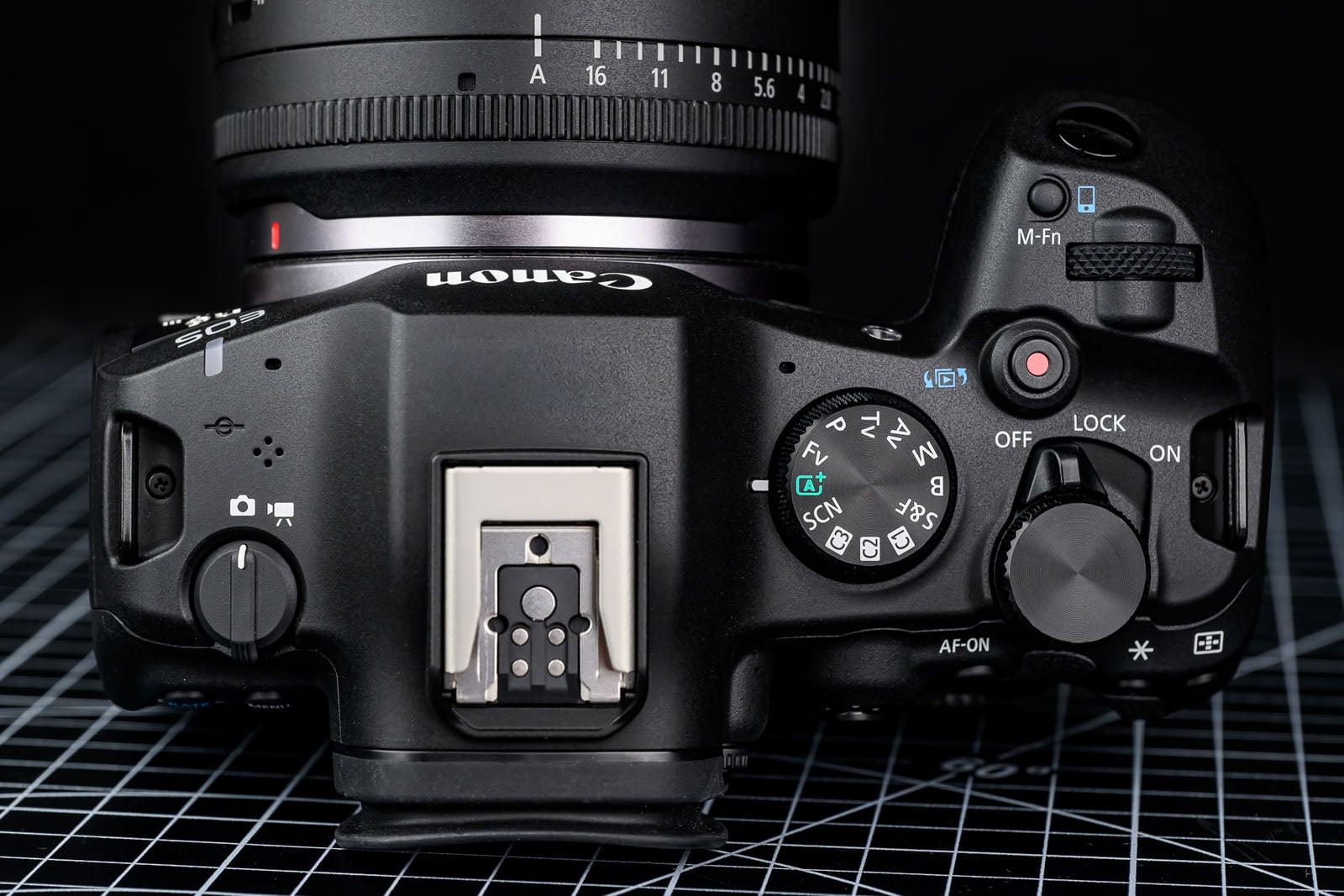 Top view of a Canon digital camera showing control dials, buttons, hot shoe mount, and part of the lens, placed on a black surface with a white grid pattern.