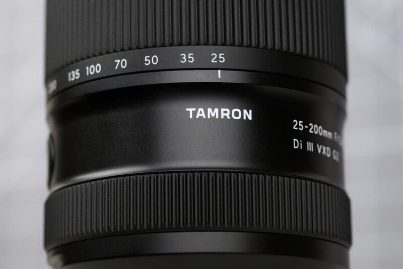 Close-up of a Tamron camera lens showing focal length markings from 25mm to 135mm, with “TAMRON” and “25-200mm Di III VXD” labeled on the lens barrel.