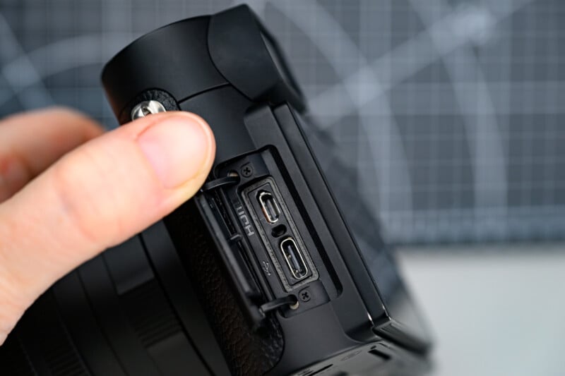 A close-up of a hand opening the side port cover of a black camera, revealing multiple ports including HDMI, USB-C, and microphone inputs. A grid-patterned surface is blurred in the background.