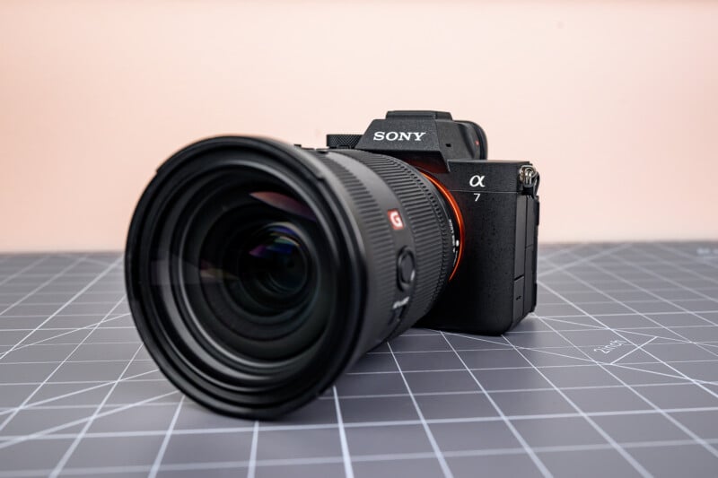 A Sony Alpha 7 camera with a large lens sits on a gray grid-patterned surface, against a light pink background.