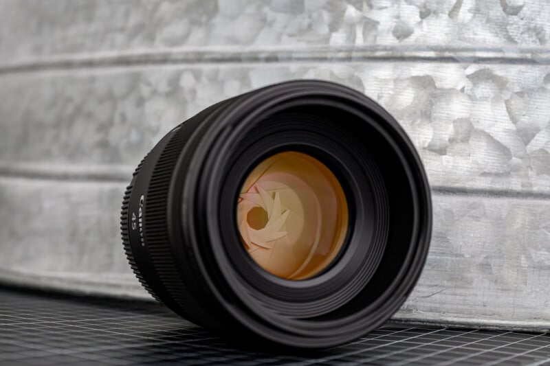 A close-up of a camera lens lying on its side on a black grid surface, with a metallic, textured background behind it. The lens glass reflects warm tones and the aperture blades are visible inside.