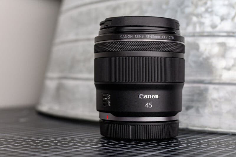 A Canon RF 45mm F1.2 STM camera lens stands upright on a black grid surface with a blurred metallic background. The Canon logo and lens details are visible on the front.
