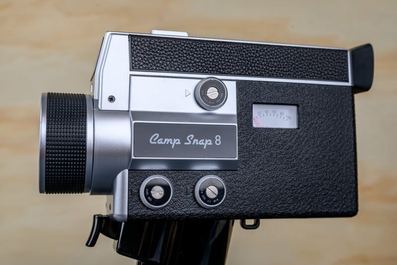 A close-up side view of a vintage black and silver Camp Snap 8 film camera with textured grip, dials, a lens, and a small gauge, set against a blurred beige background.