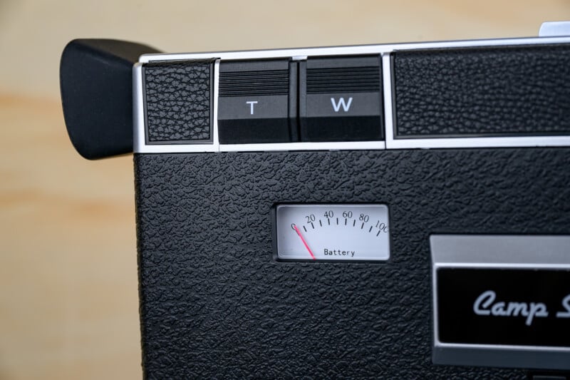 Close-up of a vintage black camera with a textured surface, showing "T" and "W" buttons, and a battery meter with the needle in the red, indicating low battery. Part of a "Camp S" logo is visible.