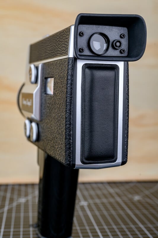 A close-up of a vintage black and silver handheld film camera with a textured grip, seen from the back and side, placed on a grid-patterned surface with a light wooden background.
