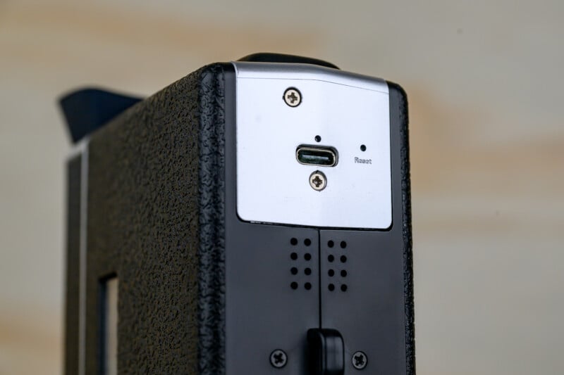 Close-up of a black and silver electronic device showing a USB-C port, a small reset button, and two screws on a textured surface. The background is out of focus.