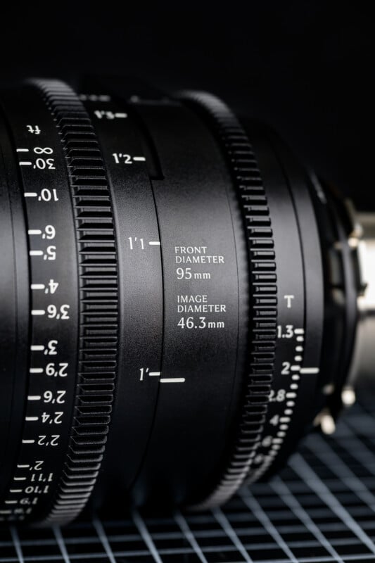 Close-up of a black camera lens showing detailed markings, including front diameter (95 mm), image diameter (46.3 mm), and focus/zoom scales, placed on a grid surface with a dark background.