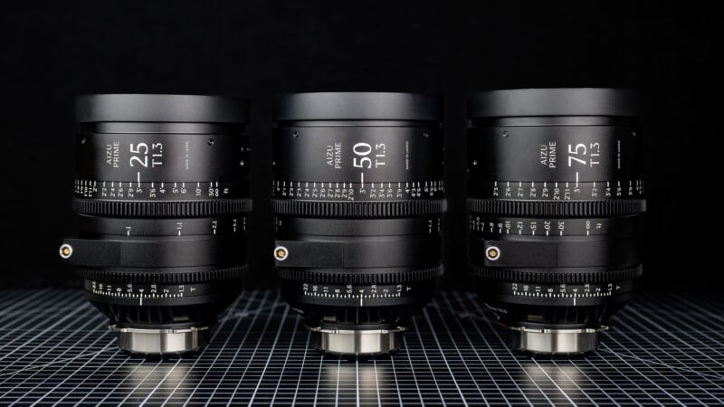Three professional camera lenses labeled 25mm, 50mm, and 75mm are lined up side by side on a checkered surface with a black background, displaying their detailed focus and aperture graduation markings.