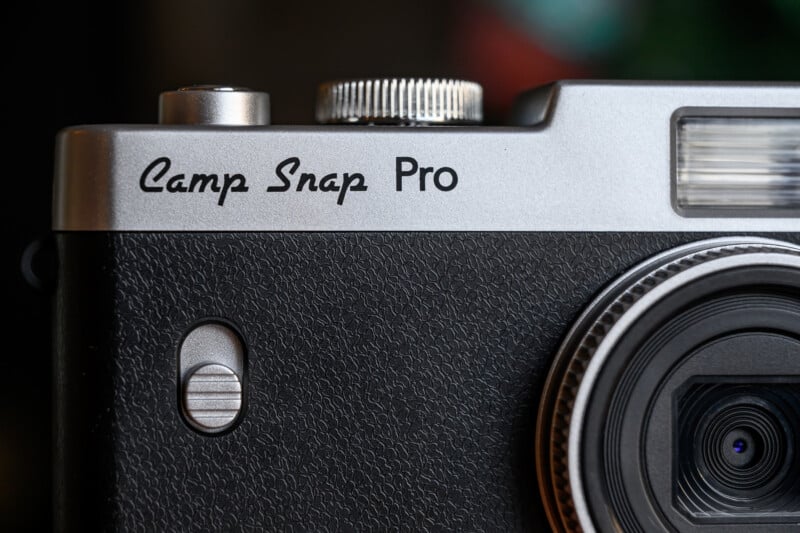 Close-up of a black and silver digital camera with the words "Camp Snap Pro" printed on the top front, showing part of the lens, flash, textured grip, and shutter button.