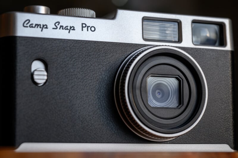 Close-up of a black and silver "Camp Snap Pro" camera showing the textured body, lens, shutter button, viewfinder, and flash. The camera is placed on a wooden surface with a blurred background.
