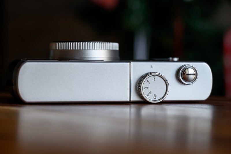 A close-up of a silver camera lying on its back, showing a textured dial, a shutter button, and a smooth metal body, with a blurred background.