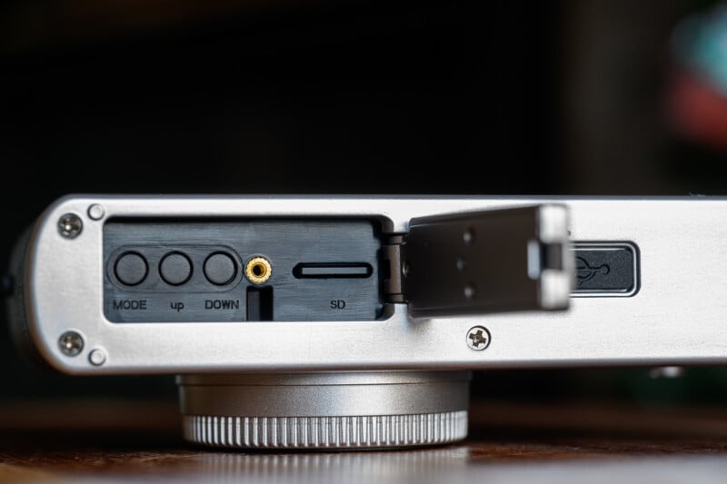 Close-up of an open port panel on an electronic device showing buttons labeled "MODE," "up," and "DOWN," a headphone jack, an SD card slot, and a USB port. The panel cover is partially open.