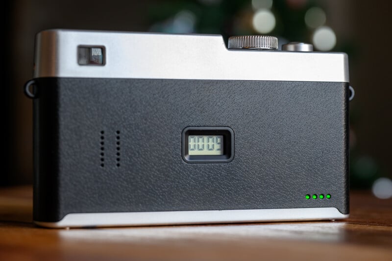 A close-up of the back of a modern film camera, showing a digital frame counter reading “0004” and three lit green indicator lights in the bottom right corner. The camera rests on a wooden surface.