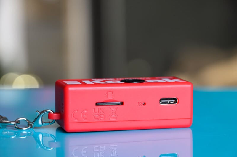 A close-up of a small red Kodak device with a keychain attached, featuring a microSD card slot, a USB-C port, and certification markings, placed on a glossy blue surface.