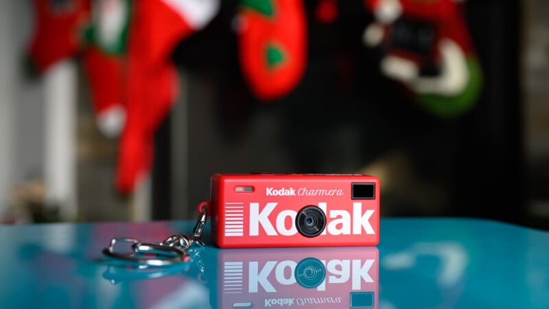 A small, red Kodak keychain camera sits on a blue surface with blurred Christmas stockings hanging in the background.