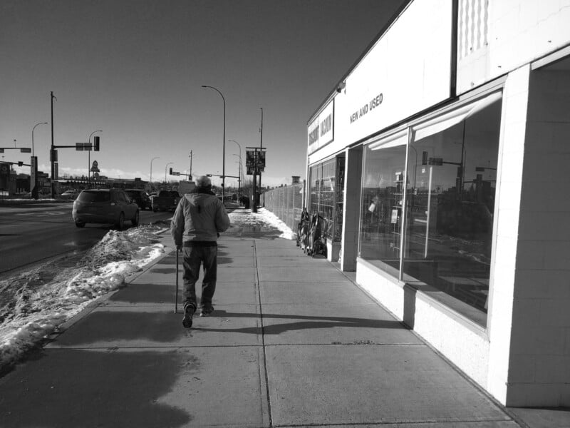 A person walks with a cane on a snowy city sidewalk beside a store with large windows, while cars drive on the adjacent road under a clear sky.