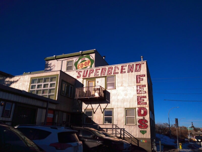 A two-story building with large red letters reading "SUPERBLEND FEEDS" on the side, a "CO-OP" sign above, and several parked cars in front under a clear blue sky.