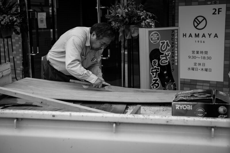 A man in a long-sleeve shirt uses a saw to cut wood on the back of a truck parked outside a shop, with Japanese signs and flowers visible in the background. The image is in black and white.