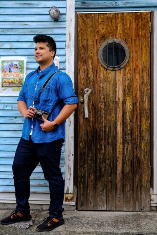 A man in a blue shirt and jeans stands holding a camera in front of a weathered wooden door with a round window, next to a blue wooden wall with posters.