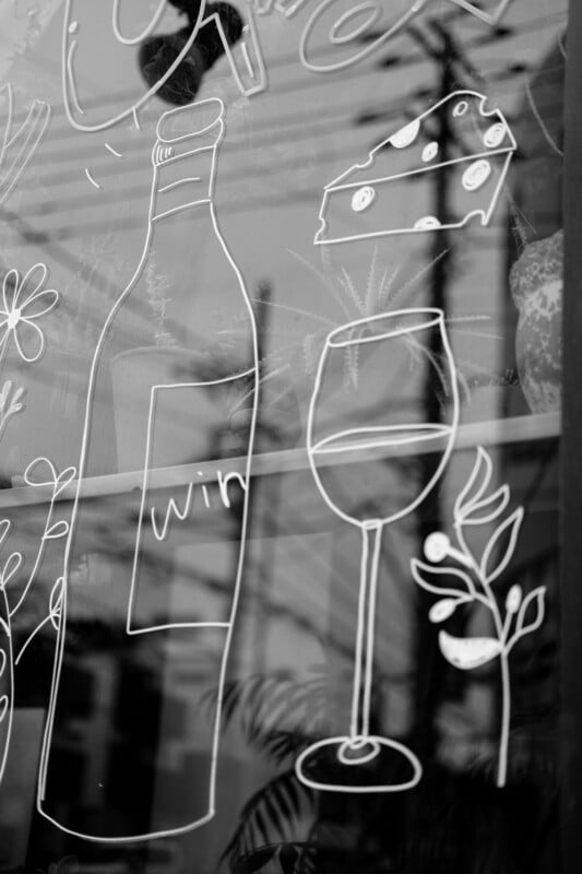 Black and white drawing on glass showing a wine bottle, a wine glass, a wedge of cheese, and some leaves, with reflections of power lines and buildings in the background.