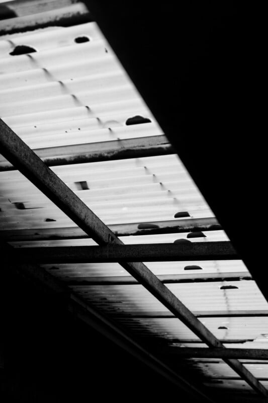 Black and white photo of a corrugated metal roof with dark spots and metal beams, creating strong shadows and a high-contrast, geometric pattern.