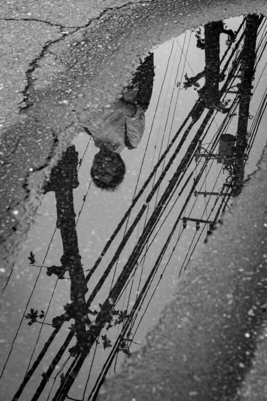 Black and white photo showing the reflection of a person and utility poles with wires in a puddle on cracked pavement. The person’s upper body and head are visible, appearing upside down in the water.