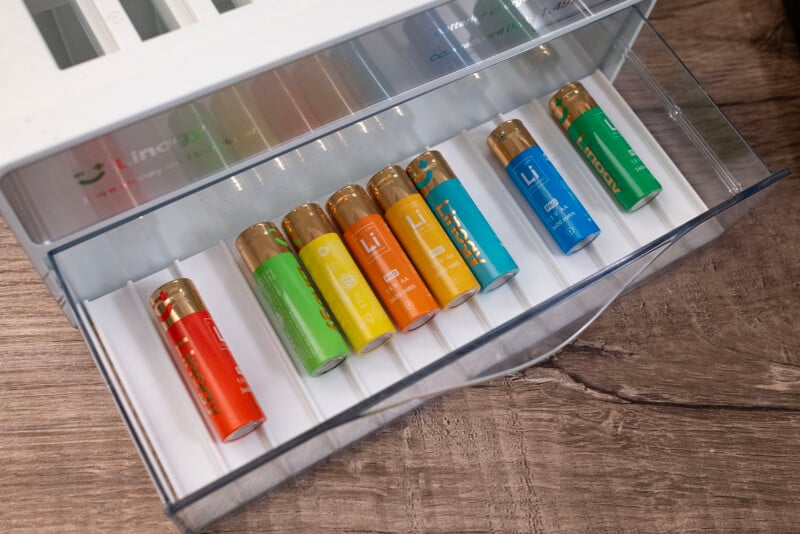 Seven rechargeable batteries in various colors (orange, red, yellow, green, blue, and teal) are organized in a white plastic drawer on a wooden surface.