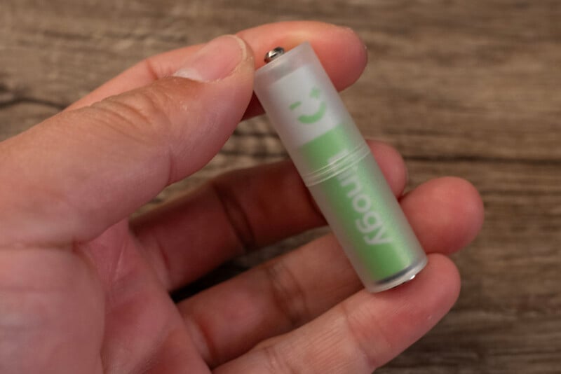 A hand holding a green rechargeable AA battery with a translucent casing, revealing the word "Imogy" and a smiling face symbol. The background is a wooden surface.