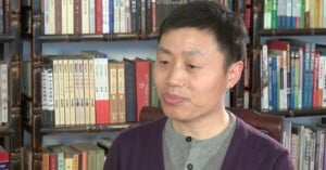 A man with short black hair and wearing a gray shirt and dark sweater sits in front of bookshelves filled with various books. He appears to be indoors and is looking slightly to the side.