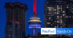 Toronto’s CN Tower is lit up in red, white, and blue at night, surrounded by tall modern buildings. The “PetaPixel Hands-On” logo appears in the lower right corner.