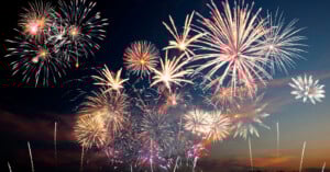 Bright, colorful fireworks burst and sparkle in the night sky over a dark landscape, creating a festive and celebratory atmosphere.