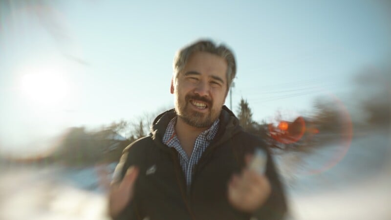 A man with gray hair and a beard smiles outdoors on a sunny winter day, wearing a dark jacket and a checkered shirt. The background is blurry with snow and bare trees, and sunlight creates a lens flare effect.